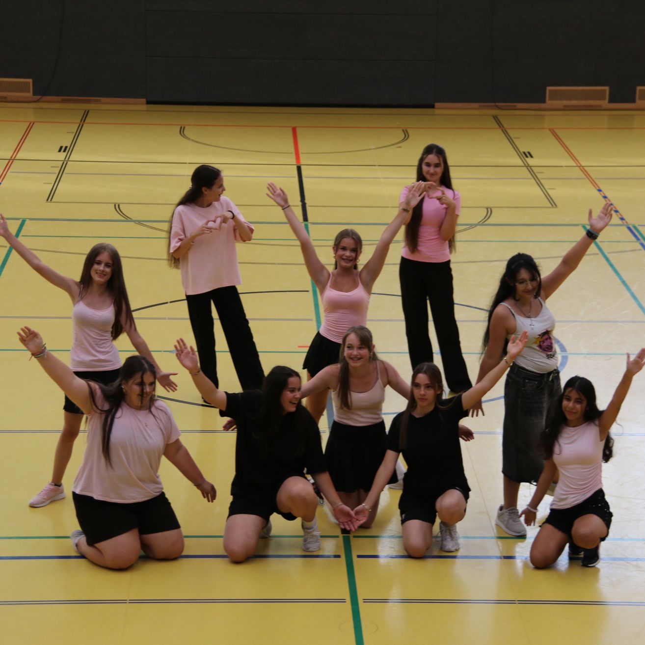 Eine Gruppe junger Frauen in passenden Outfits posiert für ein Foto auf einem Basketballplatz, einige kniend und andere stehend mit erhobenen Armen.