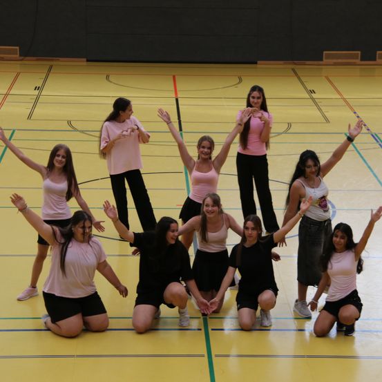 Eine Gruppe junger Frauen in passenden Outfits posiert für ein Foto auf einem Basketballplatz, einige kniend und andere stehend mit erhobenen Armen.