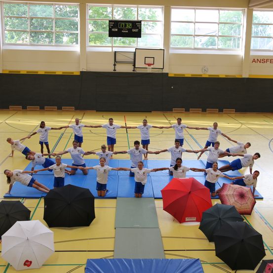 Eine Gruppe von Kindern in weißen Shirts und blauen Shorts macht Yoga-Posen auf einem blauen Teppich in einer Turnhalle. Sie halten sich an den Händen und bilden einen Kreis. Es gibt Regenschirme und Matten auf dem Boden. Die Turnhalle hat einen Basketballkorb und eine Anzeigetafel.