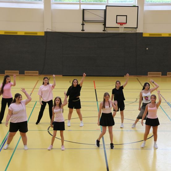 Eine Gruppe junger Frauen in verschiedenen farbigen Oberteilen und Hosen tanzt in einer Turnhalle mit einem Basketballkorb im Hintergrund.