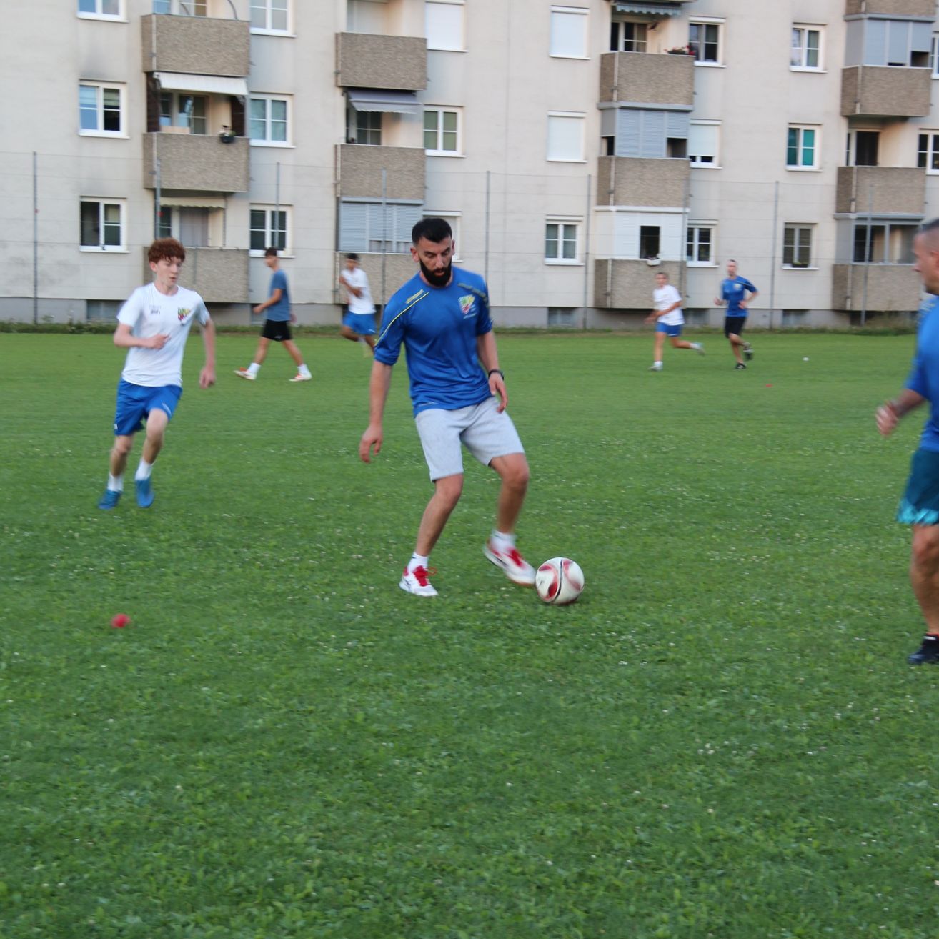 Eine Gruppe junger Männer spielt Fußball auf einem Rasenfeld, einer Mann steht kurz davor, den Ball zu treten. Sie tragen blaue und weiße Uniformen.