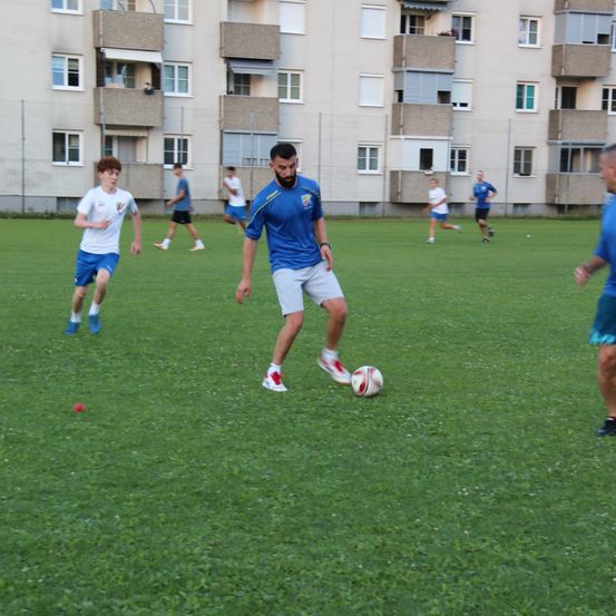 Eine Gruppe junger Männer spielt Fußball auf einem Rasenfeld, einer Mann steht kurz davor, den Ball zu treten. Sie tragen blaue und weiße Uniformen.