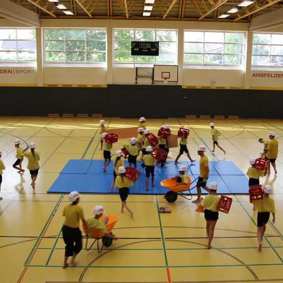 Eine Gruppe von Menschen in gelben Uniformen und weißen Helmen trägt rote Kisten und schiebt Schubkarren in einer Indoor-Sportanlage.