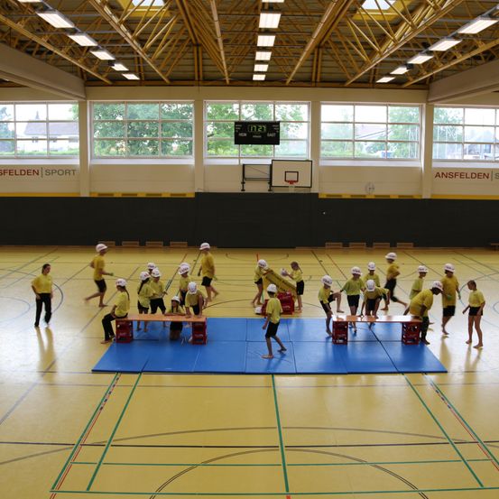 Eine Gruppe von Menschen in gelben Shirts und weißen Helmen übt eine Sportart auf einem blauen Teppich in einer Turnhalle mit einem Basketballkorb und einer Anzeigetafel.
