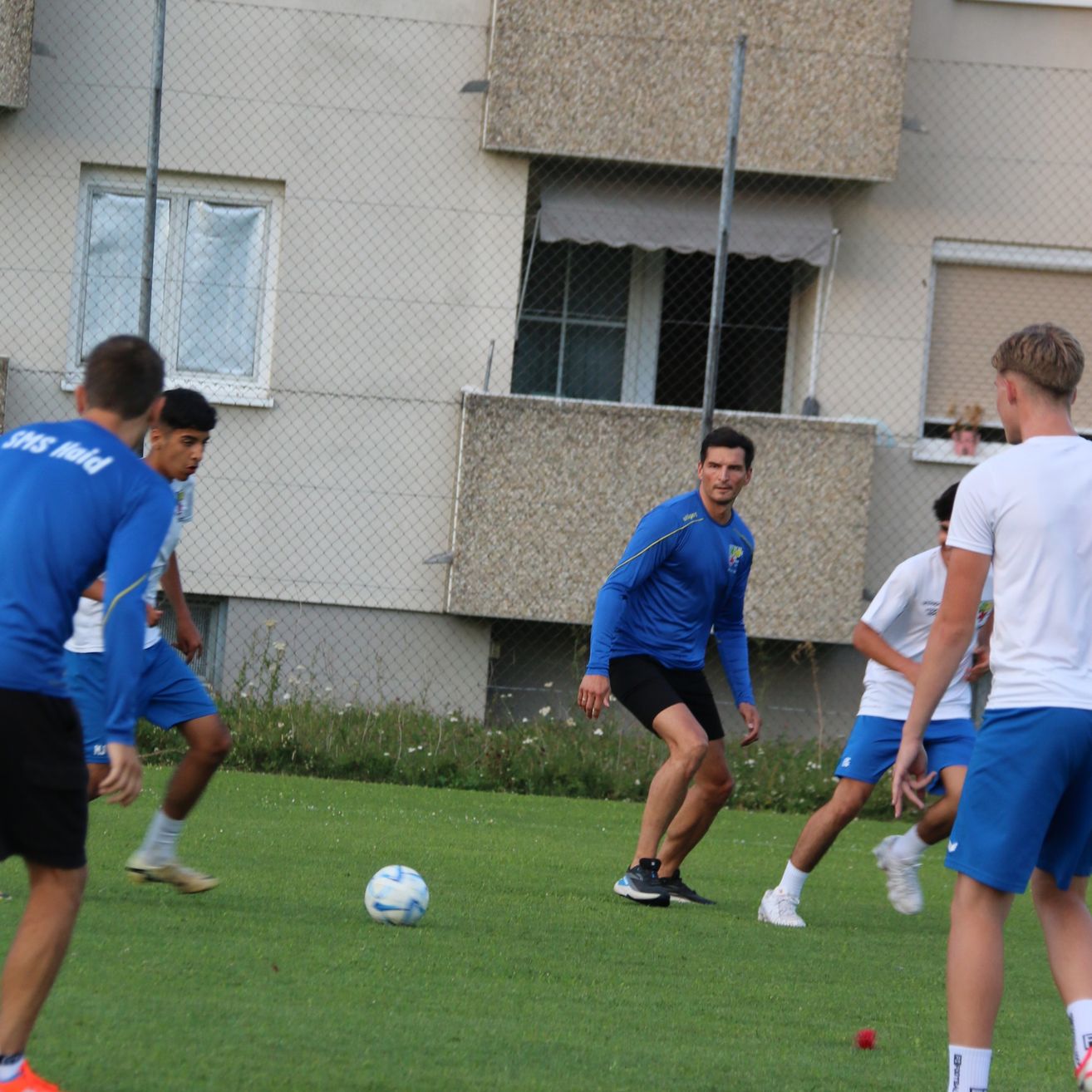 Eine Gruppe von Männern in blauen und weißen Uniformen spielt Fußball auf einem Feld mit einem Gebäude im Hintergrund.