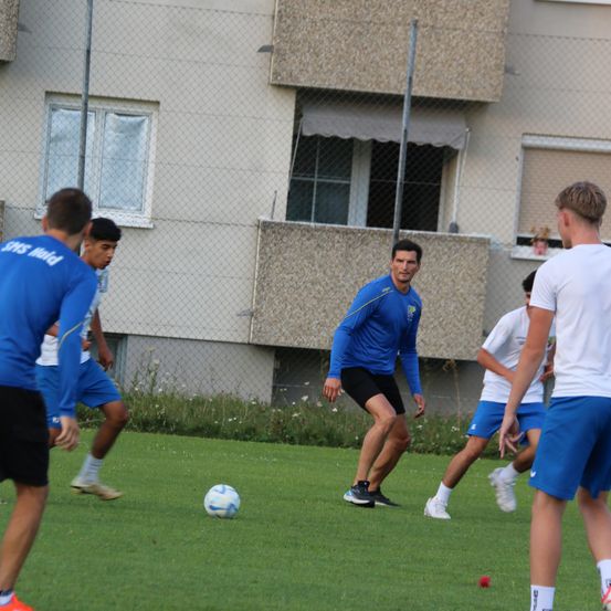 Eine Gruppe von Männern in blauen und weißen Uniformen spielt Fußball auf einem Feld mit einem Gebäude im Hintergrund.