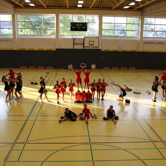 Eine Gruppe Cheerleader in roten Uniformen führt auf einem gelben Spielfeld mit einem Basketballkorb im Hintergrund eine Darbietung durch. Einige stehen auf den Schultern anderer, während andere Spagats machen.