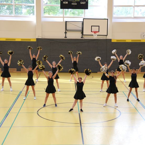 Eine Gruppe Cheerleader in schwarzen Uniformen führt auf einem Hallenbasketballplatz auf, hält Pom-Poms und lächelt. Der Platz hat gelbe Markierungen und einen Basketballkorb im Hintergrund.