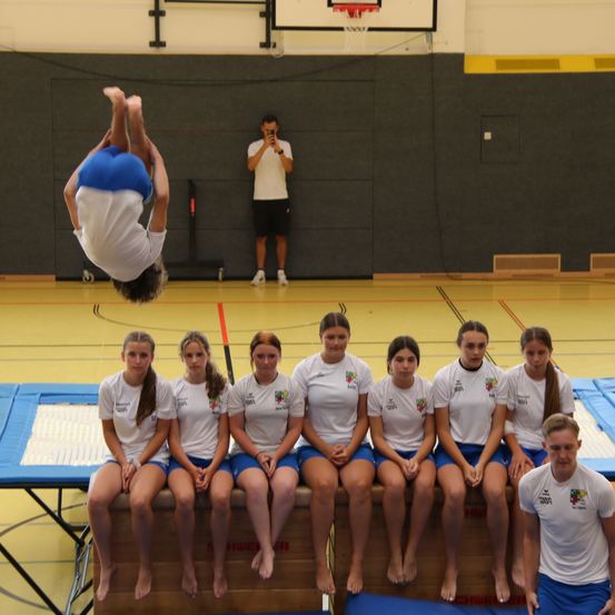 Eine Gruppe junger Frauen in weißen T-Shirts und blauen Shorts sitzt auf einer Holzkiste in einer Turnhalle. Ein Mann in einem weißen Shirt macht einen Handstand in der Luft. Ein anderer Mann steht hinter ihnen und macht ein Foto.