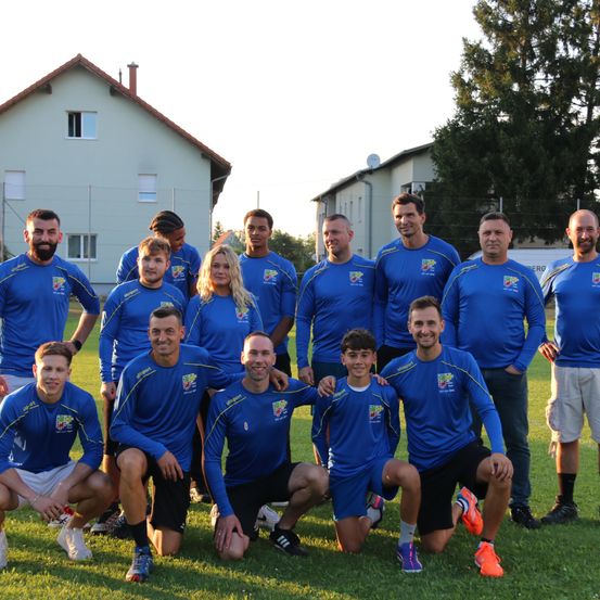 Eine Gruppe von Fußballspielern in blauen Trikots posiert für ein Foto auf einem Rasenfeld mit Häusern im Hintergrund.