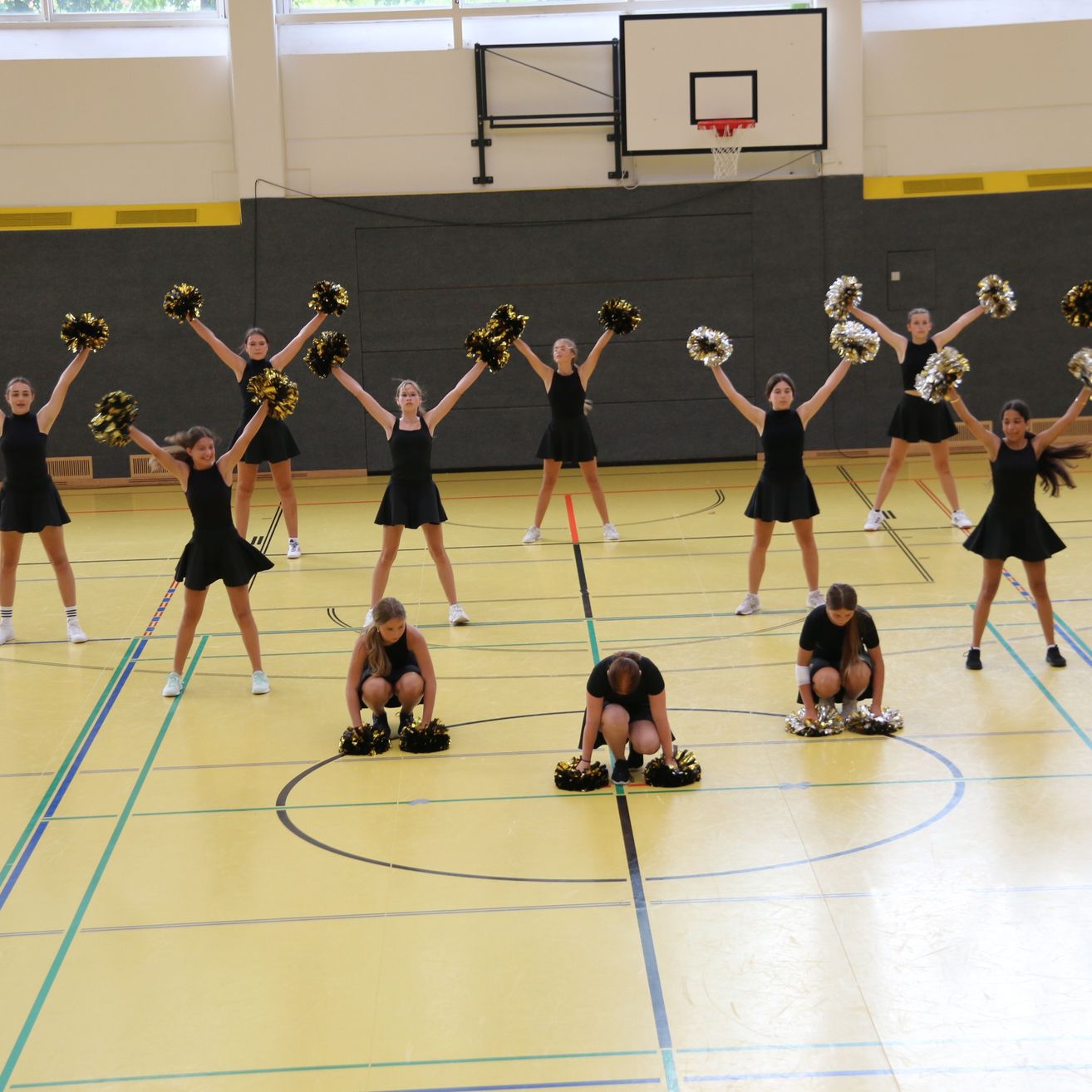 Eine Gruppe Cheerleader in schwarzen Uniformen führt auf einem gelben Spielfeld mit blauen und grünen Markierungen auf. Einige knien, während andere mit Pom-Poms stehen. Ein Basketballkorb ist im Hintergrund sichtbar.