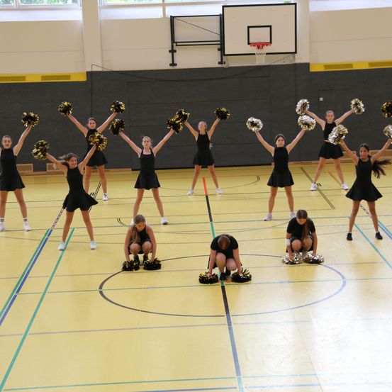 Eine Gruppe Cheerleader in schwarzen Uniformen führt auf einem gelben Spielfeld mit blauen und grünen Markierungen auf. Einige knien, während andere mit Pom-Poms stehen. Ein Basketballkorb ist im Hintergrund sichtbar.