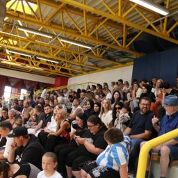 Eine große Menschenmenge sitzt in einem Stadion und beobachtet eine Veranstaltung. Einige tragen Brillen, Hüte und Turnschuhe. Das Stadion hat gelbe Stützen und blaue Wände.