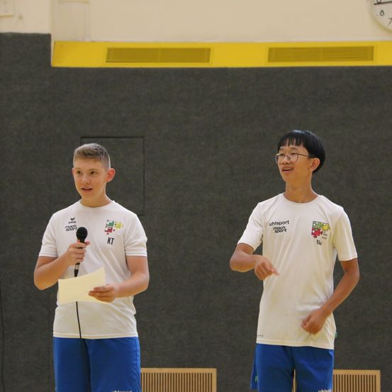 Zwei junge Jungen stehen in einer Turnhalle, einer hält ein Mikrofon und der andere gestikuliert. Sie tragen beide weiße T-Shirts mit Logos und blaue Shorts. Hinter ihnen befindet sich eine graue Wand mit einem gelben Rand.
