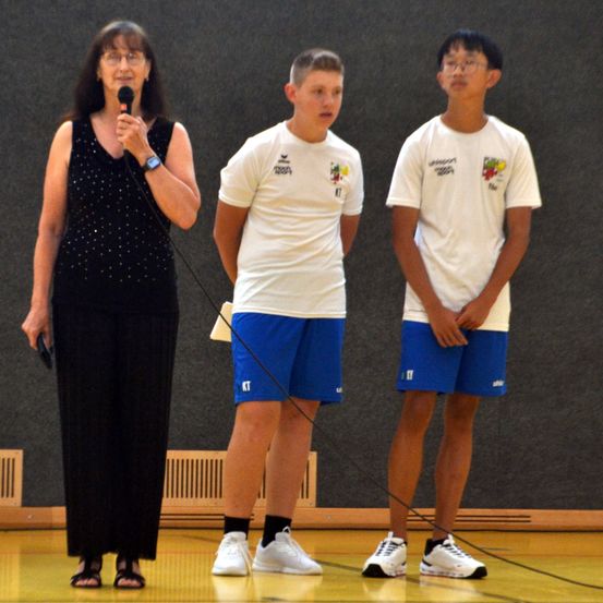 Eine Frau mit einem Mikrofon steht zwischen zwei Jungen in Sportkleidung, möglicherweise bei einer Preisverleihung. Die Jungen tragen weiße T-Shirts mit blauen Shorts und Turnschuhen.