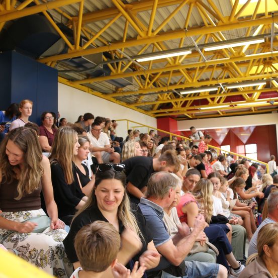 Eine große Menschenmenge sitzt in einem Stadion, einige lächeln und klatschen. Das Stadion hat gelbe Stützen und rote Wände.