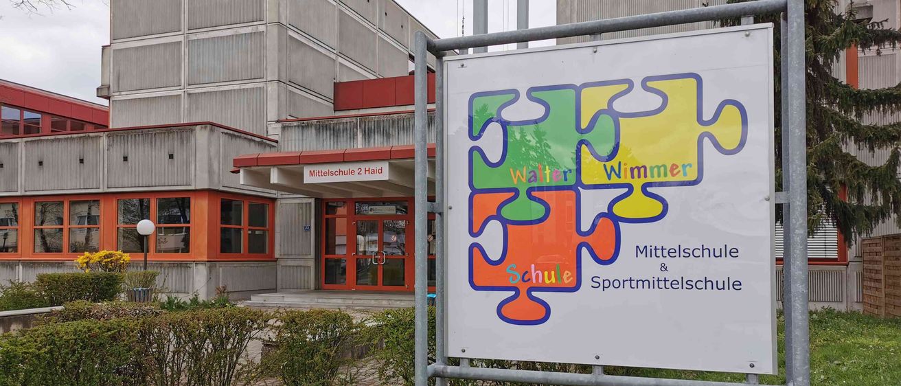Ein Schulschild für die Walter Wimmer Mittel- und Sportschule steht vor einem Gebäude. Das Gebäude hat ein rotes Dach und Glastüren.