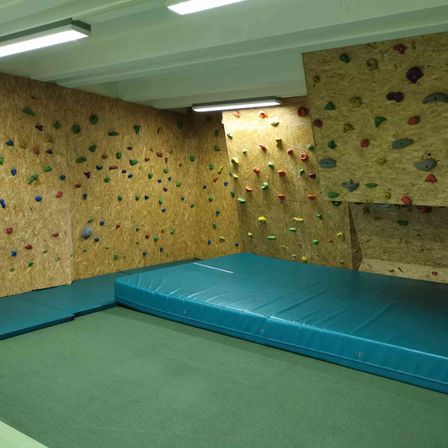 Ein Indoor-Kletterraum mit einer blauen Matratze auf dem Boden und Kletterwänden, die mit farbigen Griffen bedeckt sind.