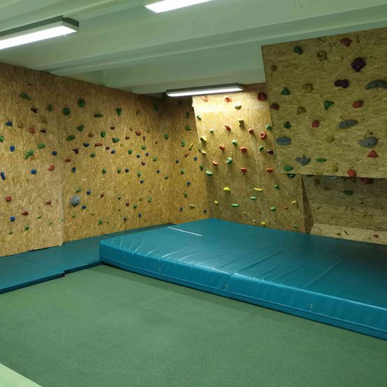 Ein Indoor-Kletterraum mit einer blauen Matratze auf dem Boden und Kletterwänden, die mit farbigen Griffen bedeckt sind.