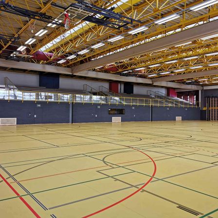 Ein Hallenbasketballplatz mit gelbem Boden und roten, grünen und blauen Markierungen. Der Platz ist von leeren Tribünen und einem gelben Geländer umgeben. Die Decke hat gelbe Metallrahmen und weiße Lampen.