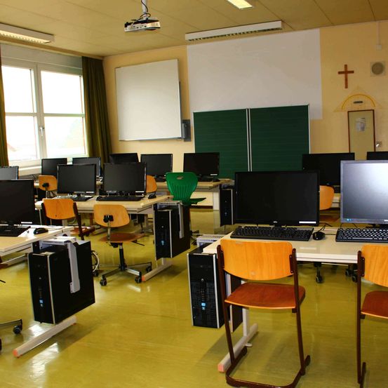 Ein Klassenzimmer mit mehreren Computerbildschirmen und Tastaturen auf Tischen, einem Projektor darüber und einem Kreuz an der Wand.