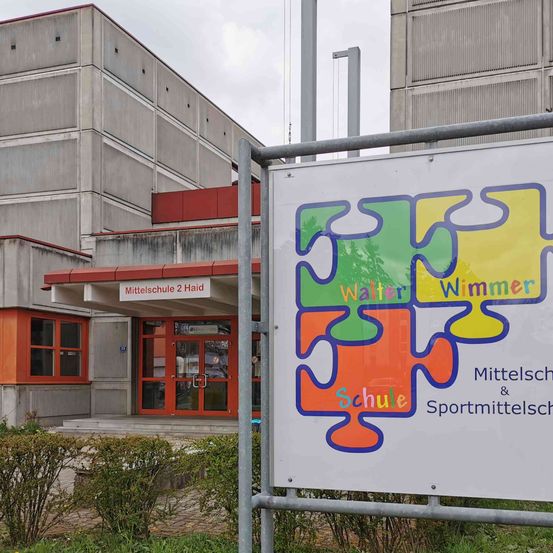 Ein Schild vor dem Walter Wimmer Mittelschule 2 Gebäude mit Puzzleteilen, dem Wort 'Schule' und 'Sportmittelschule'. Hinter dem Schild steht ein Schulgebäude mit roten Akzenten.