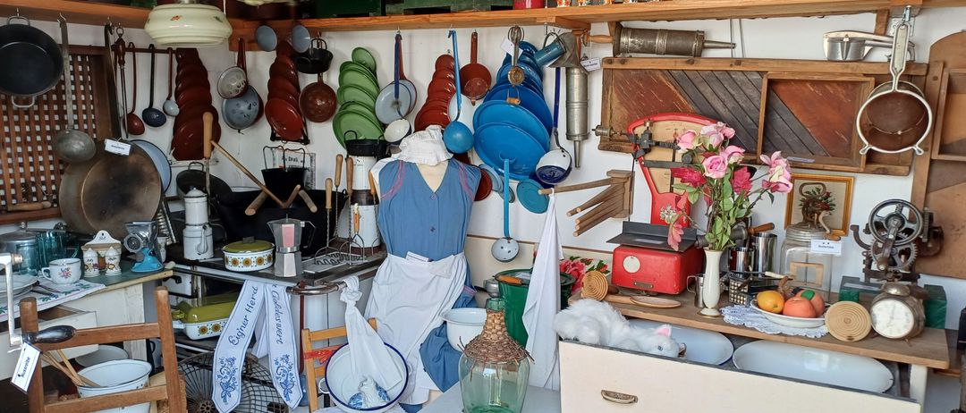 Eine Küche, gefüllt mit Vintage-Töpfen, Pfannen und Küchenutensilien zur Schau gestellt. Ein Mannequin in einem blauen Kleid steht zwischen den Gegenständen. Regale halten verschiedene Küchenwerkzeuge und Dekorationen. Ein kleiner Tisch und ein Stuhl sind in der Nähe, mit einem Glas und Blumen auf der Arbeitsplatte.