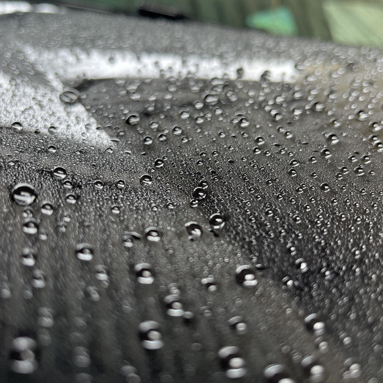 Eine Nahaufnahme von zahlreichen Wassertropfen auf einer schwarzen Oberfläche, möglicherweise der Windschutzscheibe eines Autos. Die Tropfen sind verstreut, einige überlappen sich.