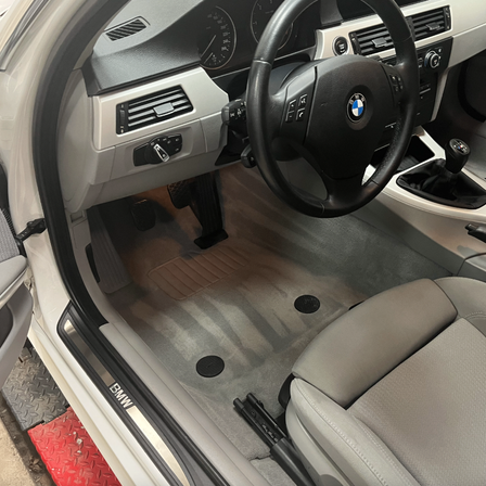 Das Innere eines BMW-Fahrzeugs ist zu sehen, wobei die Fahrertür geöffnet ist. Das Lenkrad und das Armaturenbrett sind sichtbar, zusammen mit einem Gangschalter und Pedalen. Die Fußmatten sind entfernt, und das graue Interieur des Autos ist zu sehen.