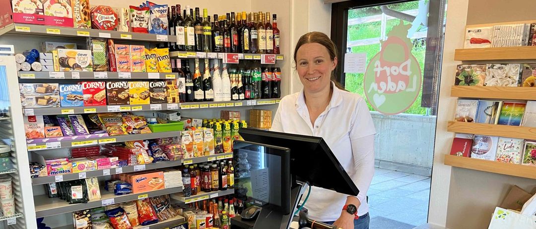 Eine Frau steht lächelnd hinter einer Theke in einem Geschäft. Hinter ihr befinden sich Regale mit verschiedenen Snacks und Getränken. Auf dem Tresen sind ein Monitor, eine Kasse und andere Geräte. Rechts von ihr befindet sich eine Glastür mit einem Logo.