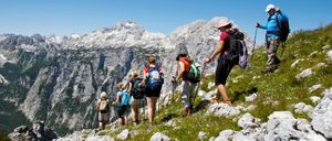 Eine Gruppe von Wanderern steigt einen grasbewachsenen Berghang mit schneebedeckten Gipfeln im Hintergrund hinauf. Sie sind mit Rucksäcken und Trekkingstöcken ausgestattet.