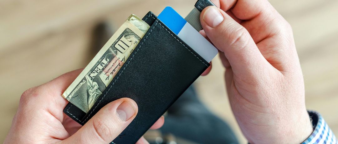 Eine Hand hält ein schwarzes Portemonnaie mit einer blauen Karte und einem Dollarschein sichtbar. Das Portemonnaie scheint ein schlankes, minimalistisches Design zu sein, möglicherweise aus Leder.