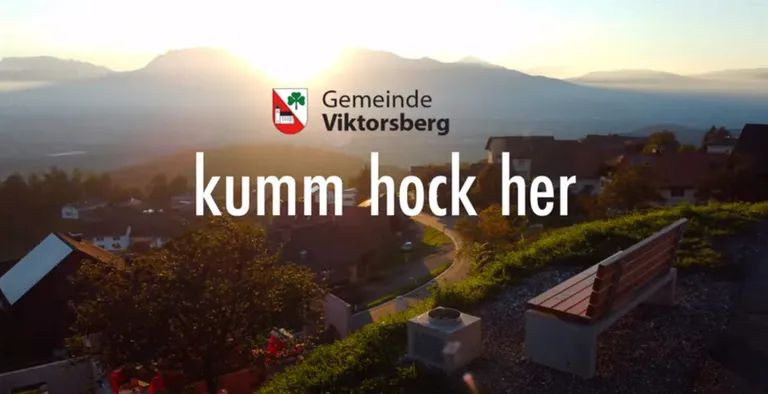 Eine Landschaft von Viktorsberg mit einer Bank, Grünflächen und Bergen unter dem untergehenden Sonnenlicht. Der Text lautet 'kumm hock her'.