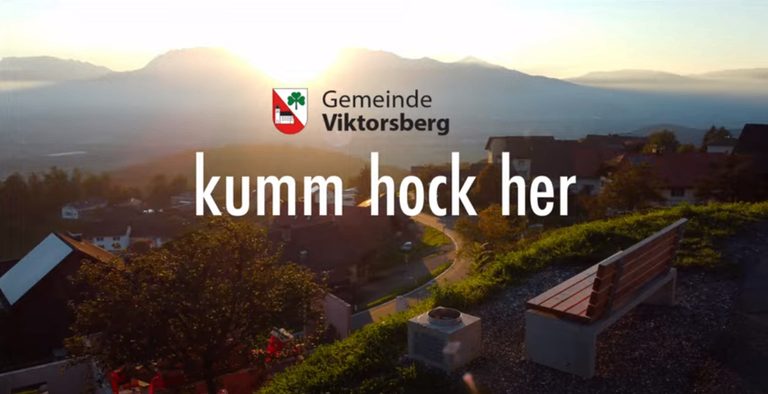 Eine Landschaft von Viktorsberg mit einer Bank, Grünflächen und Bergen unter dem untergehenden Sonnenlicht. Der Text lautet 'kumm hock her'.