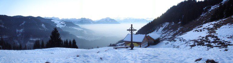 Ein hölzernes Kreuz steht im Schnee vor einer Hütte mit schneebedecktem Dach. Das Gebiet ist von Bäumen umgeben, und Berge sind in der Ferne mit Nebel.