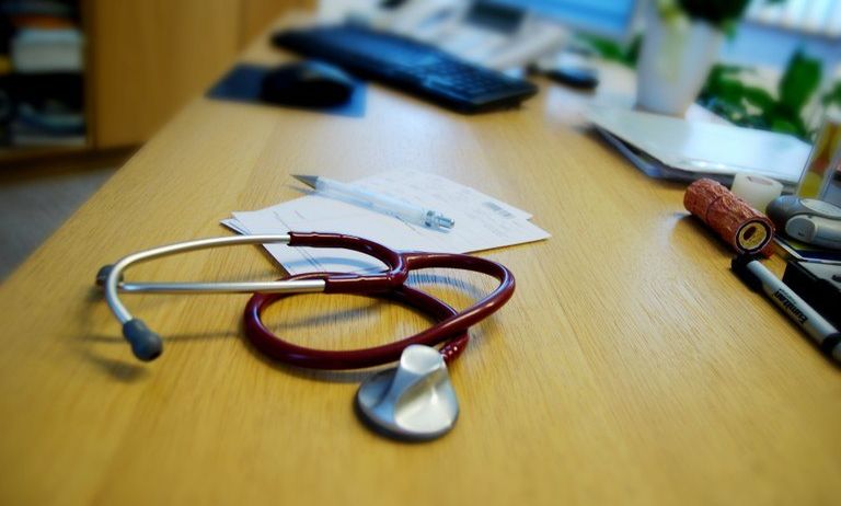 Ein Arztschreibtisch mit Stethoskop, Stift, Notizblock, Tastatur und Topfpflanze auf einem Regal.