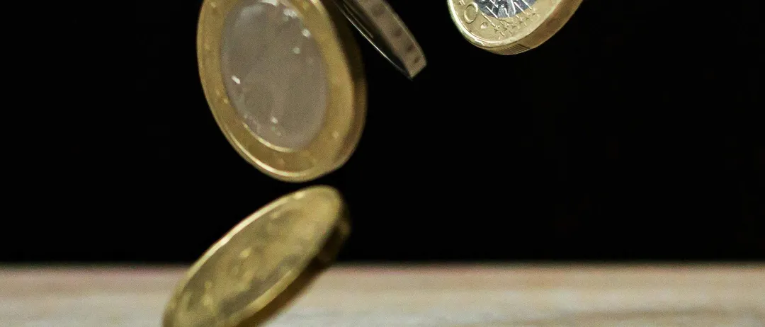 Vier Münzen befinden sich in der Luft, jede in einem anderen Stadium des Überschlagens. Sie sind goldfarben und haben komplizierte Designs. Der Hintergrund ist dunkel, mit einer hölzernen Oberfläche darunter.
