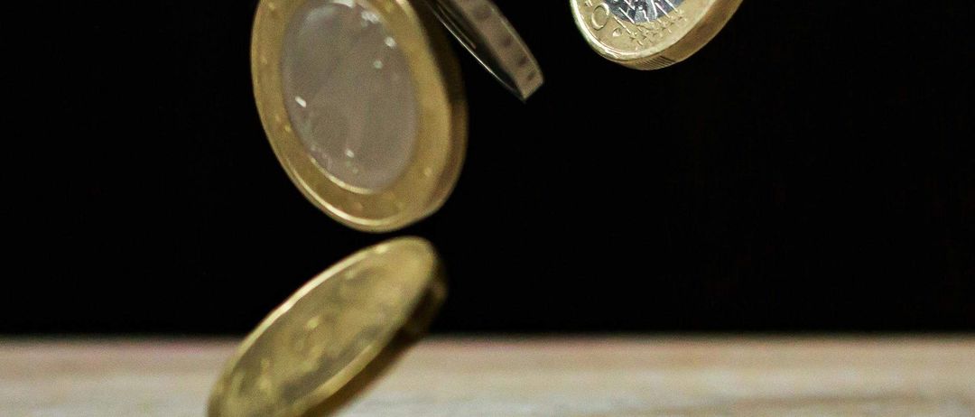 Vier Münzen befinden sich in der Luft, jede in einem anderen Stadium des Überschlagens. Sie sind goldfarben und haben komplizierte Designs. Der Hintergrund ist dunkel, mit einer hölzernen Oberfläche darunter.