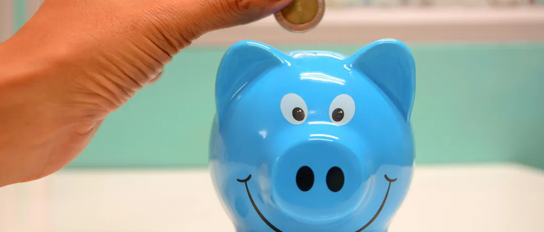 Eine Hand legt eine Münze in eine lächelnde blaue Sparschwein auf einer weißen Oberfläche.