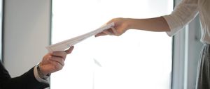 Eine Person in formeller Kleidung überreicht ein Dokument an eine andere Person in einem weißen Hemd in einer Büroumgebung.