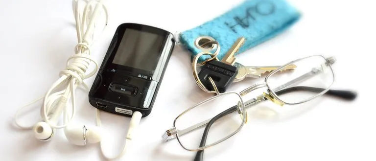 Ein schwarzer MP3-Player, weiße Kopfhörer, eine Brille, ein blauer Schlüsselanhänger und zwei Schlüssel liegen auf einer weißen Oberfläche.
