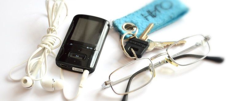 Ein schwarzer MP3-Player, weiße Kopfhörer, eine Brille, ein blauer Schlüsselanhänger und zwei Schlüssel liegen auf einer weißen Oberfläche.