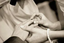 Ein Bräutigam und eine Braut tauschen während einer Hochzeitszeremonie Ringe aus, die ihr Engagement füreinander symbolisieren. Die Hand des Bräutigams hält einen Ring, während die Braut ihn an seinen Finger legt. Beide tragen Eheringe, und die Braut trägt eine Perlenkette.