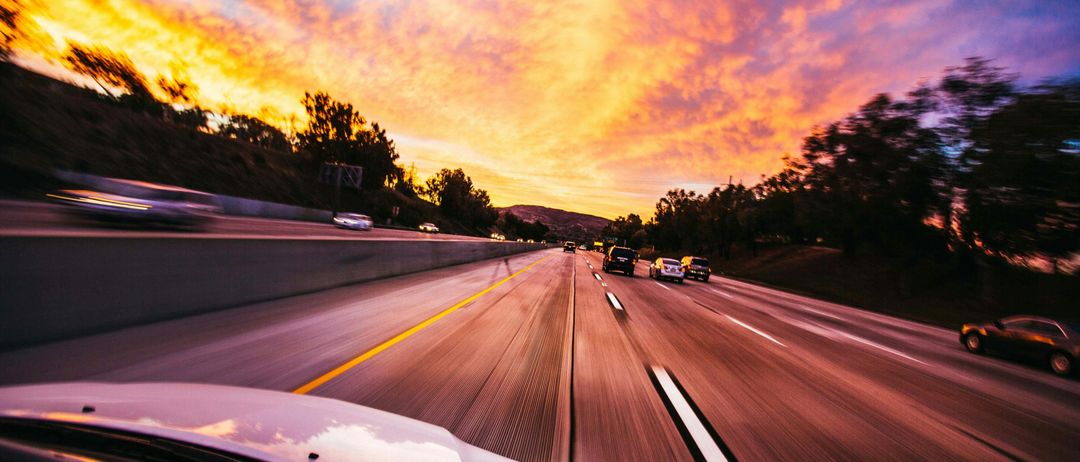 Autobahnansicht mit Sonnenuntergangsfarben, schnell fahrende Autos, Bäume an den Seiten und klarer Himmel.