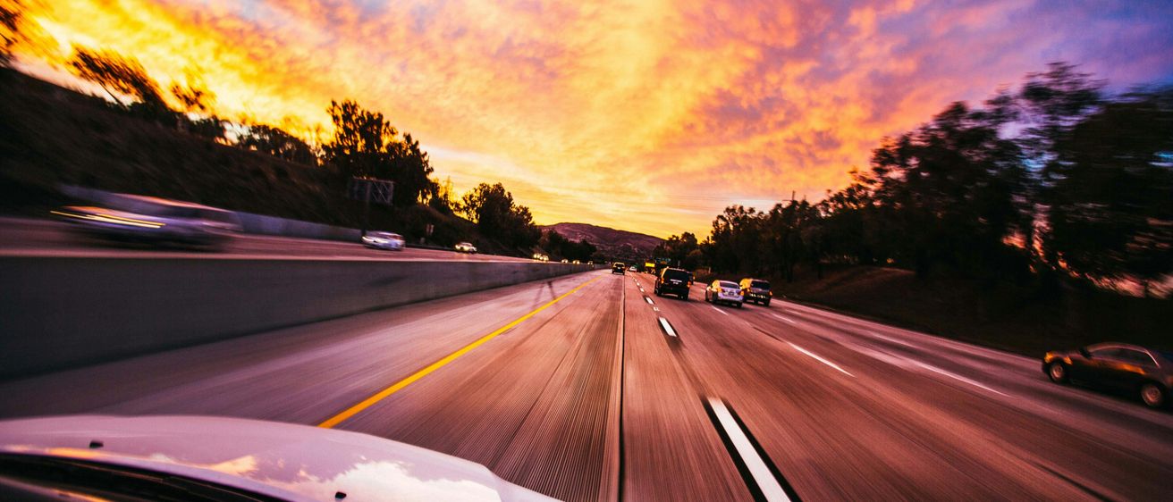 Autobahnansicht mit Sonnenuntergangsfarben, schnell fahrende Autos, Bäume an den Seiten und klarer Himmel.