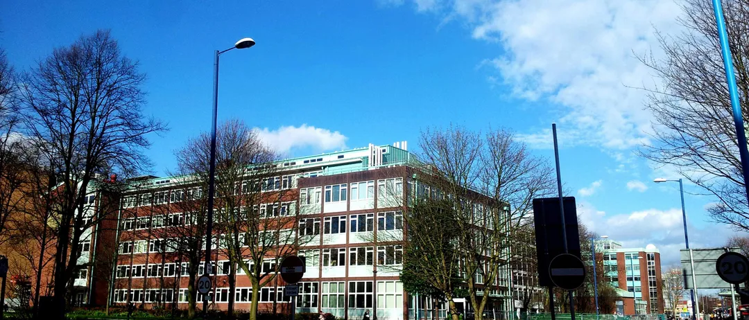 Ein mehrstöckiges Gebäude mit vielen Fenstern steht unter einem blauen Himmel mit Wolken. Straßenlaternen säumen den Bürgersteig. Eine Person geht die Straße entlang.