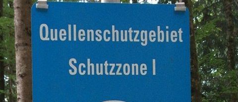 Ein blaues Schild, das auf ein Quellschutzgebiet hinweist, gekennzeichnet als Schutzzone I. Das Schild hat ein blaues und weißes kreisförmiges Symbol mit Wellenlinien.