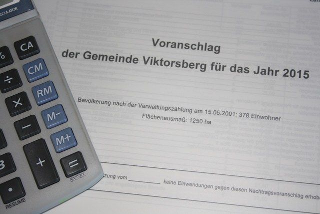 Ein Taschenrechner liegt auf einem Dokument mit dem Titel 'Vorschlag der Gemeinde Viktorsberg fur das Jahr 2015'. Das Dokument enthält Details wie Bevölkerungszahl und Landfläche.