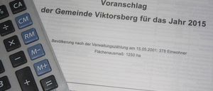 Ein Taschenrechner liegt auf einem Dokument mit dem Titel 'Vorschlag der Gemeinde Viktorsberg fur das Jahr 2015'. Das Dokument enthält Details wie Bevölkerungszahl und Landfläche.