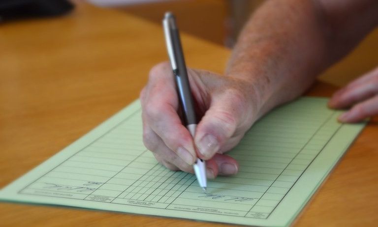 Eine ältere Person schreibt mit einem Stift auf einem Holztisch auf ein Formular. Das Formular hat mehrere Zeilen.